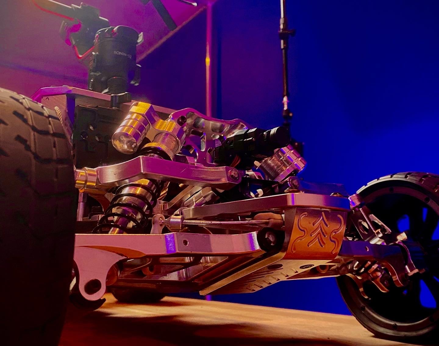 Close-up of an aluminum Baja RC chassis and suspension under purple and blue workshop lighting.
